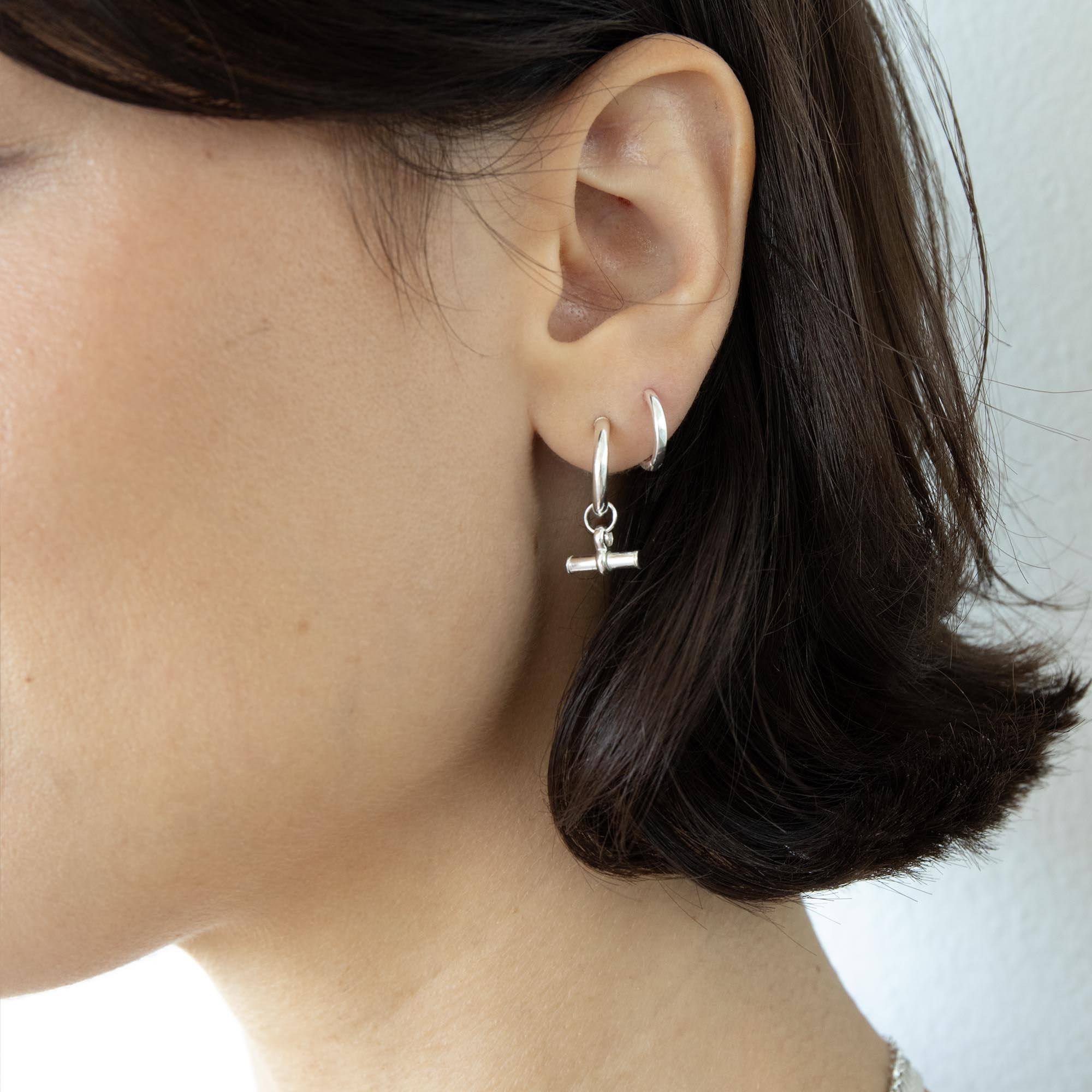 A close-up of a person's ear wearing a silver huggie hoop earring with a small dangling barbell charm - seolgold
