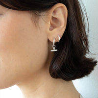 A close-up of a person's ear wearing a silver huggie hoop earring with a small dangling barbell charm - seolgold