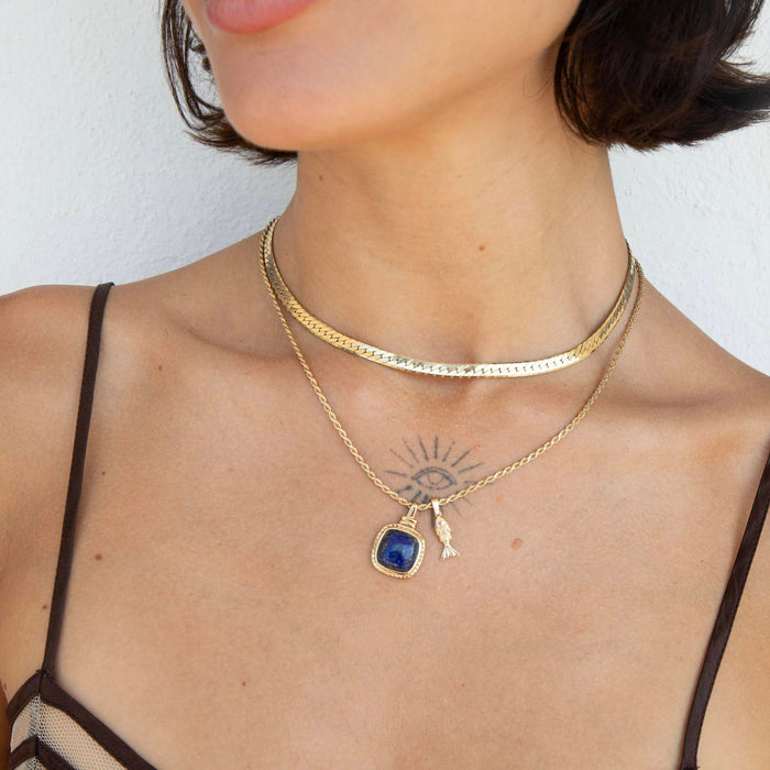 A close-up of a model wearing a layered gold necklace set. The top layer is a textured gold choker, and the bottom layer is a delicate gold chain featuring a square-cut dark blue pendant and a small gold key charm - seolgold