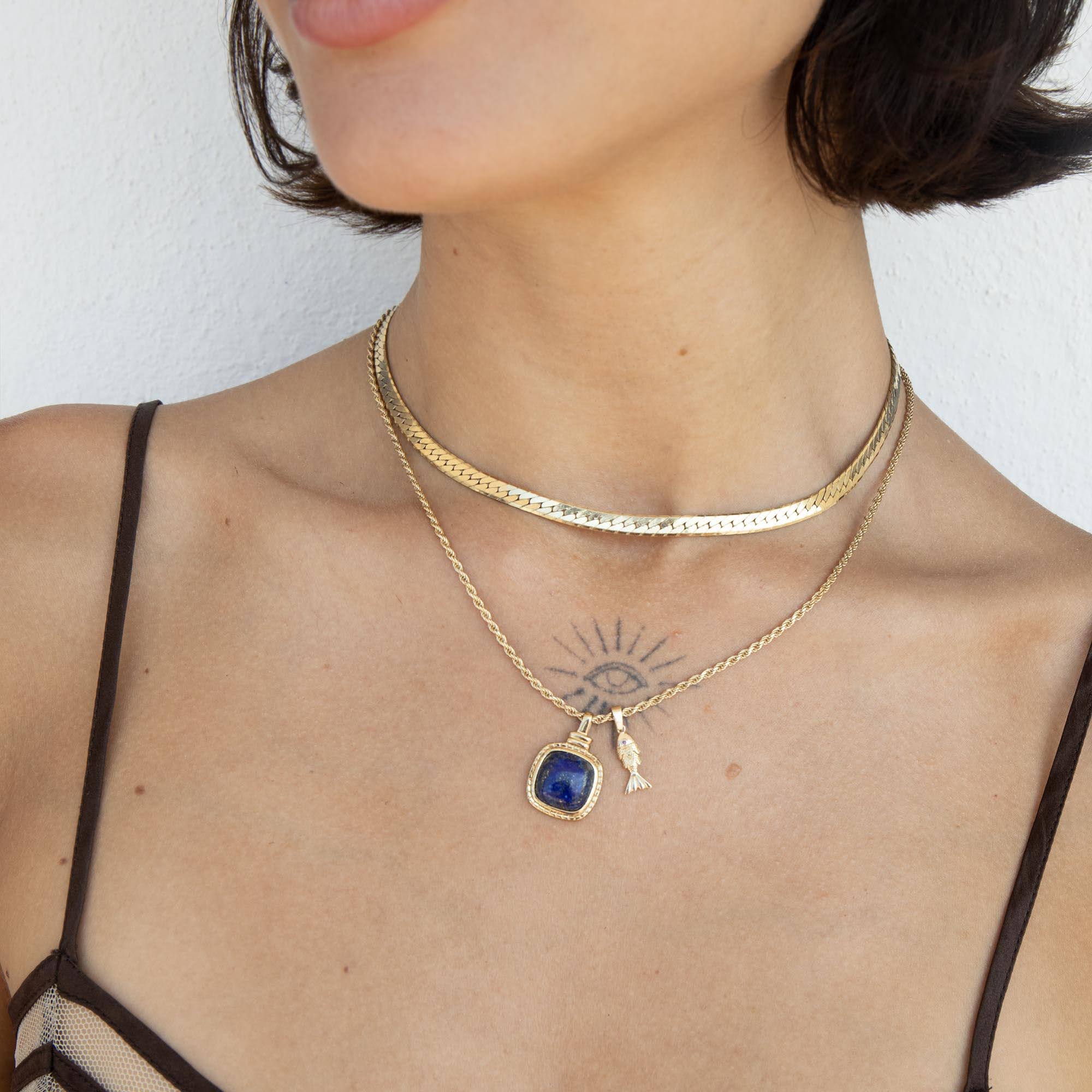 A close-up of a model wearing a layered gold necklace set. The top layer is a textured gold choker, and the bottom layer is a delicate gold chain featuring a square-cut dark blue pendant and a small gold key charm - seolgold