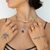 A close-up lifestyle shot of a model wearing layered gold necklaces, including a textured choker, a delicate chain with a sun pendant, and a chain with a blue oval pendant, along with several gold rings - seolgold