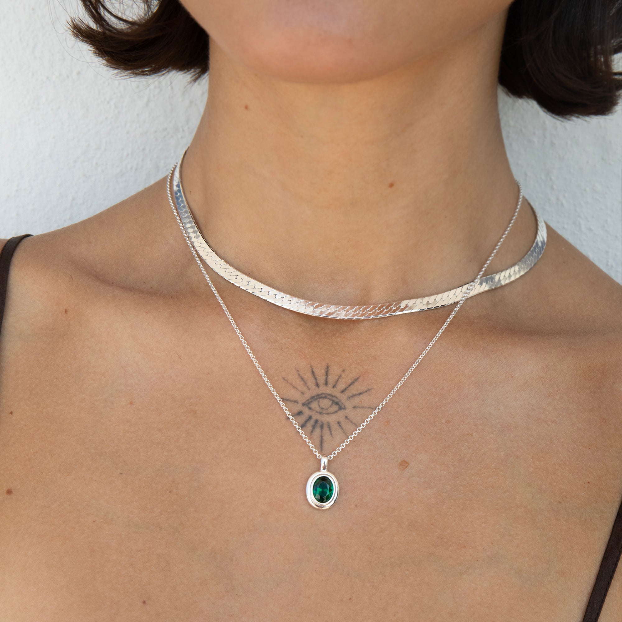 A close-up of a model's neck and chest wearing two layered silver necklaces: a textured choker and a delicate pendant necklace with a green emerald oval stone - seolgold