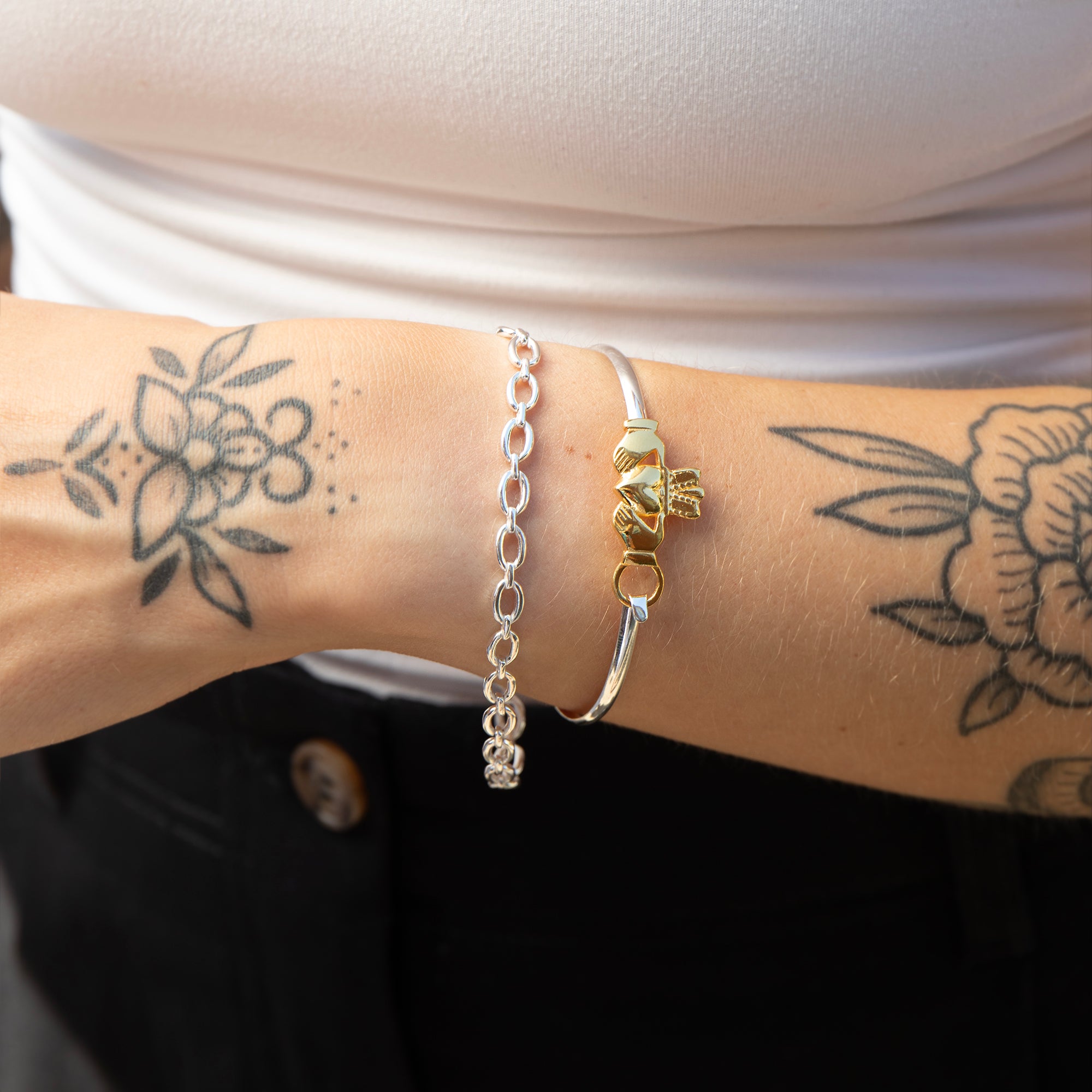 A close-up of a person's tattooed wrist wearing two bracelets: a silver chain bracelet and a silver bangle with a gold-toned claddagh motif. Seol gold