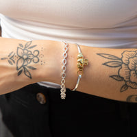 A close-up of a person's tattooed wrist wearing two bracelets: a silver chain bracelet and a silver bangle with a gold-toned claddagh motif. Seol gold