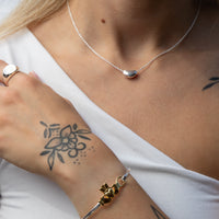 A close-up lifestyle shot of a woman wearing a delicate silver pendant necklace, a silver ring, and a gold-and-silver geometric bangle bracelet, styled with a white top. Seol Gold