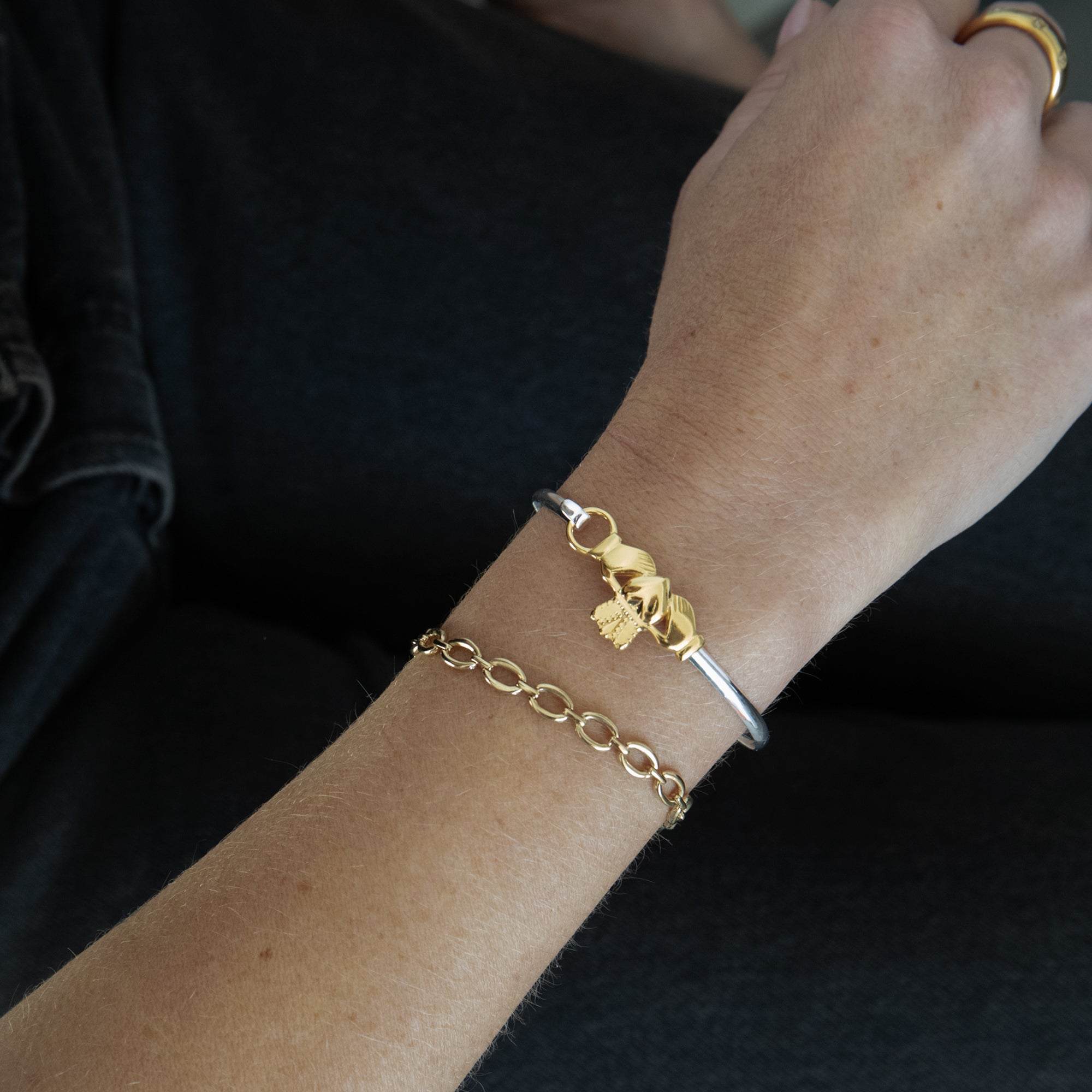 A close-up of a person's wrist wearing two delicate bracelets: a gold chain bracelet and a silver bangle with a gold harp charm, against a dark navy shirt background - seol gold