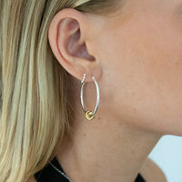 A close-up of a woman's ear wearing a large silver hoop earring with a small gold heart knot detail. Seol gold