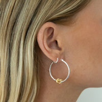 A close-up of a model's ear wearing a pair of silver hoop earrings with a small gold knot detail and tiny clear stones at the top. Seol Gold
