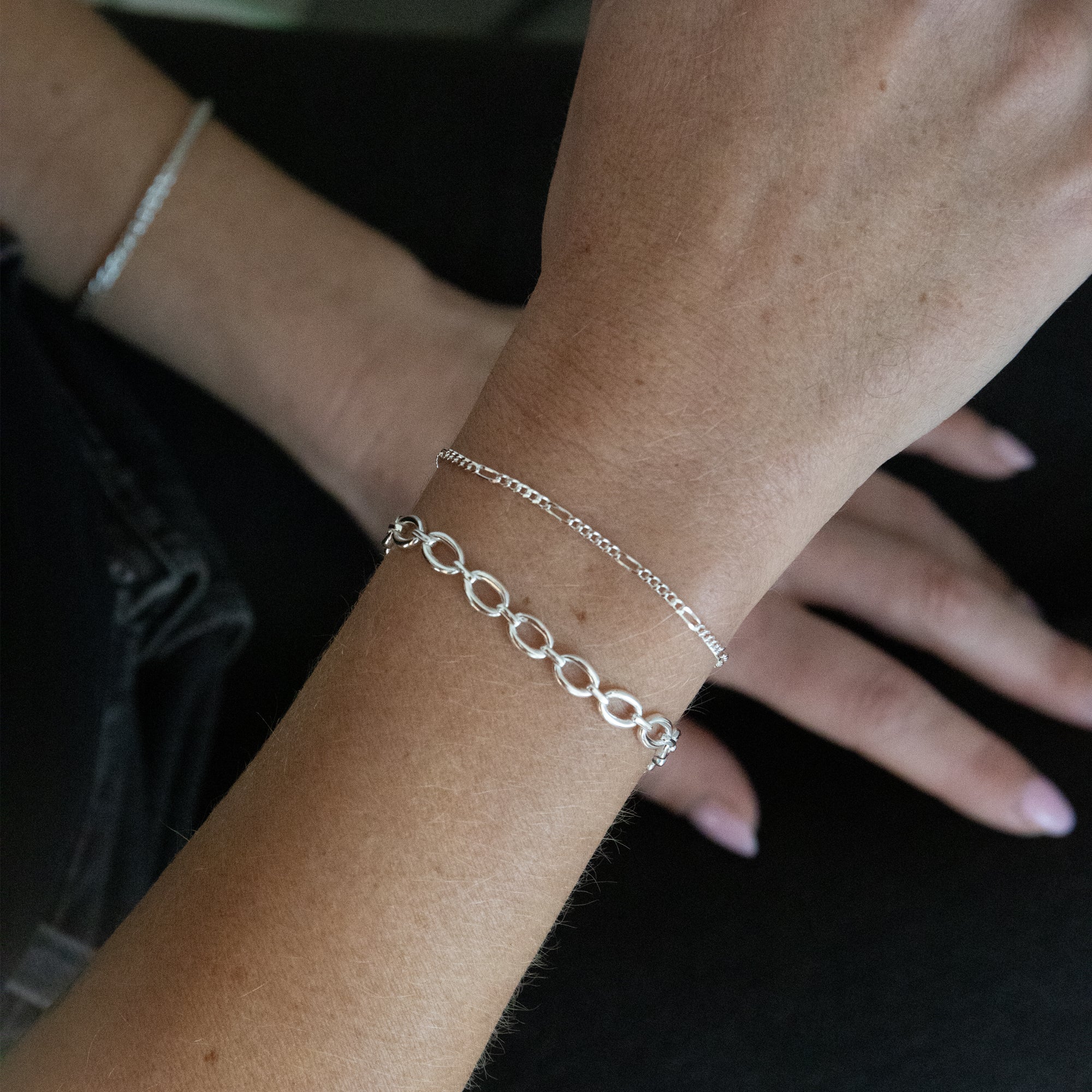 A close-up of a person's wrist wearing two delicate silver bracelets, with one bracelet featuring interlocking oval links and the other a fine curb chain, against a dark background. Seol Gold