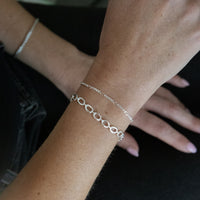 A close-up of a person's wrist wearing two delicate silver bracelets, with one bracelet featuring interlocking oval links and the other a fine curb chain, against a dark background. Seol Gold