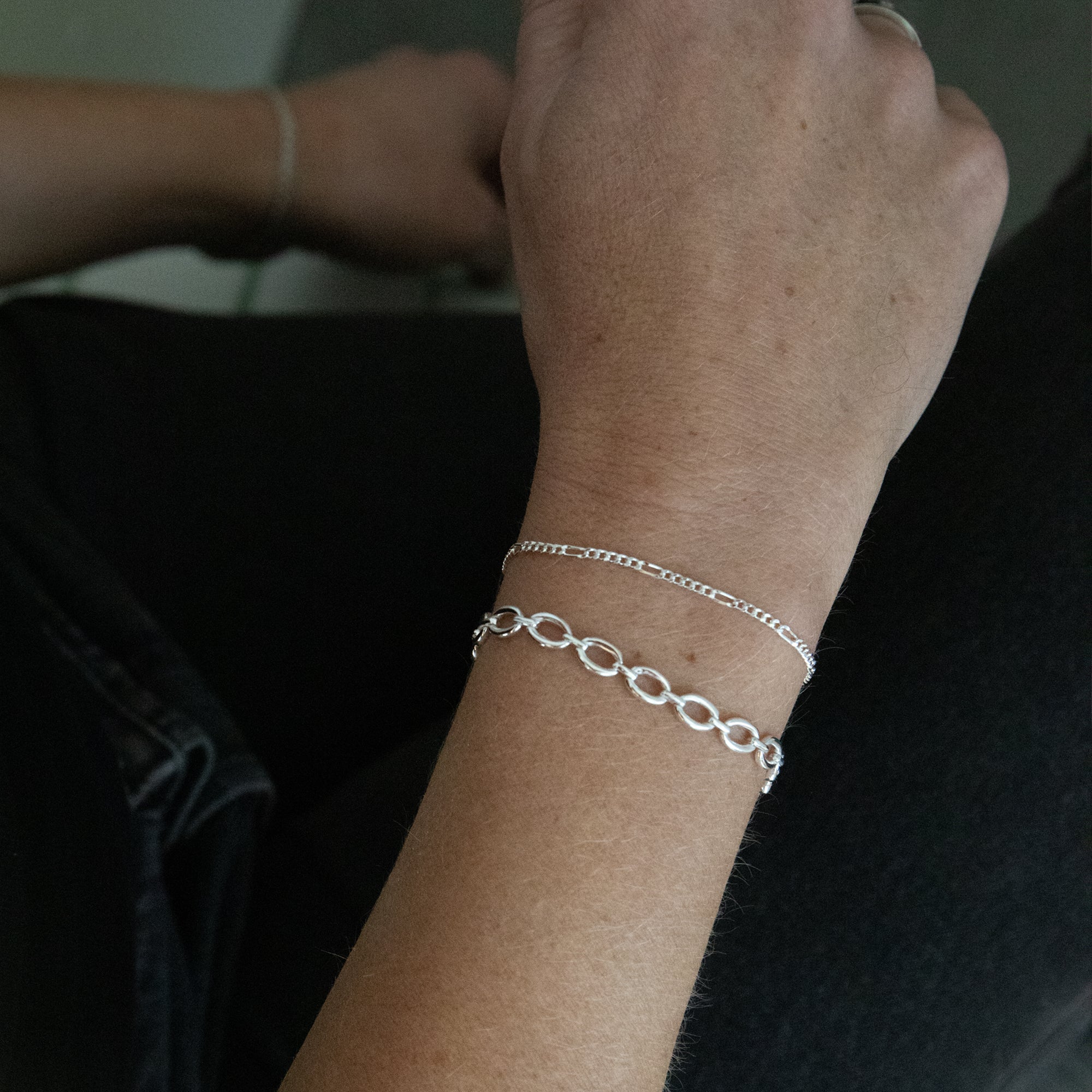 A close-up of a person's wrist wearing two delicate silver bracelets, one a fine chain and the other an oval-link chain, against a dark background. seol gold