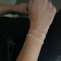 A close-up of a person's wrist wearing two delicate silver bracelets, one a fine chain and the other an oval-link chain, against a dark background. seol gold