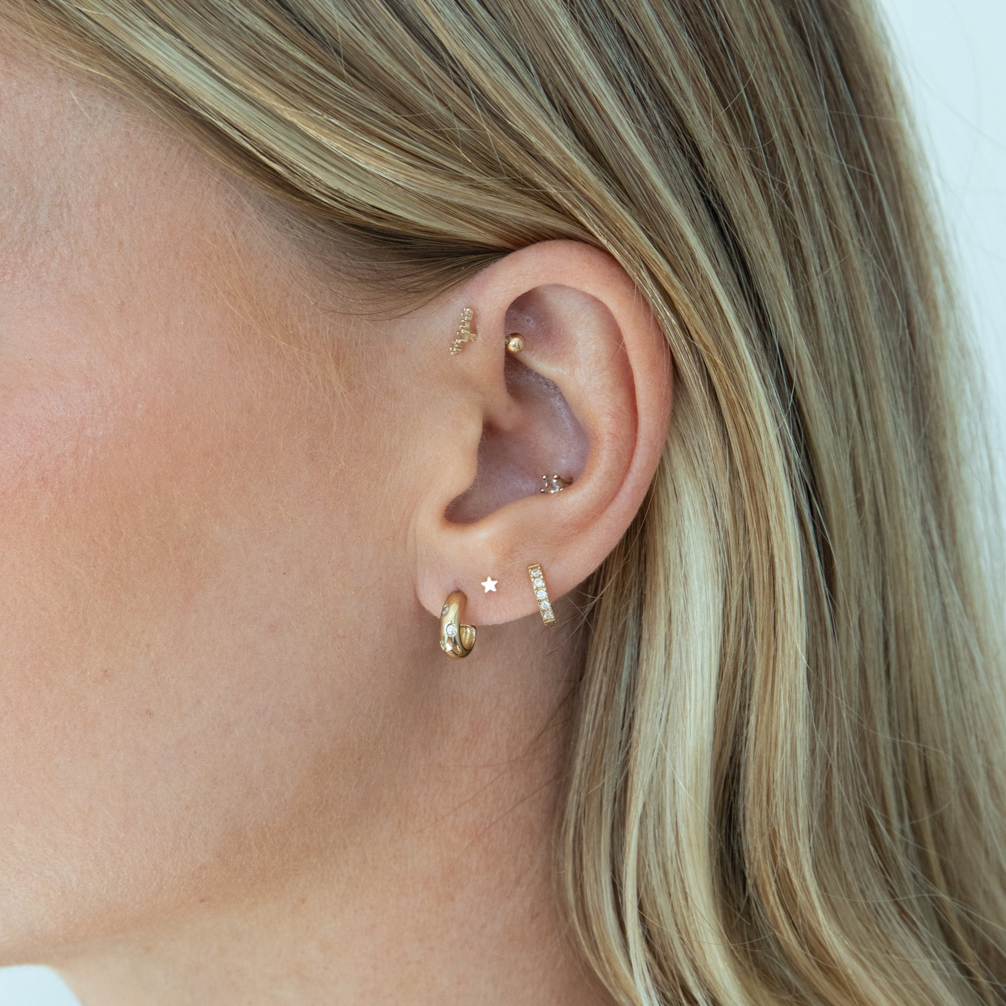 Close-up of a person's ear wearing multiple gold ear piercings and earrings, including hoop, stud, and cartilage piercings, against a light neutral background - seolgold