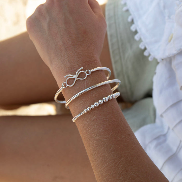 Close-up of a wrist wearing multiple silver bracelets with a blurred background - silver bow bangle - seolgold