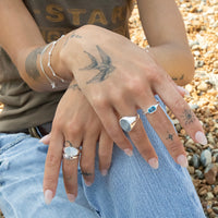 Close-up of hands with tattoos wearing silver rings on a pebbly surface - seolgold
