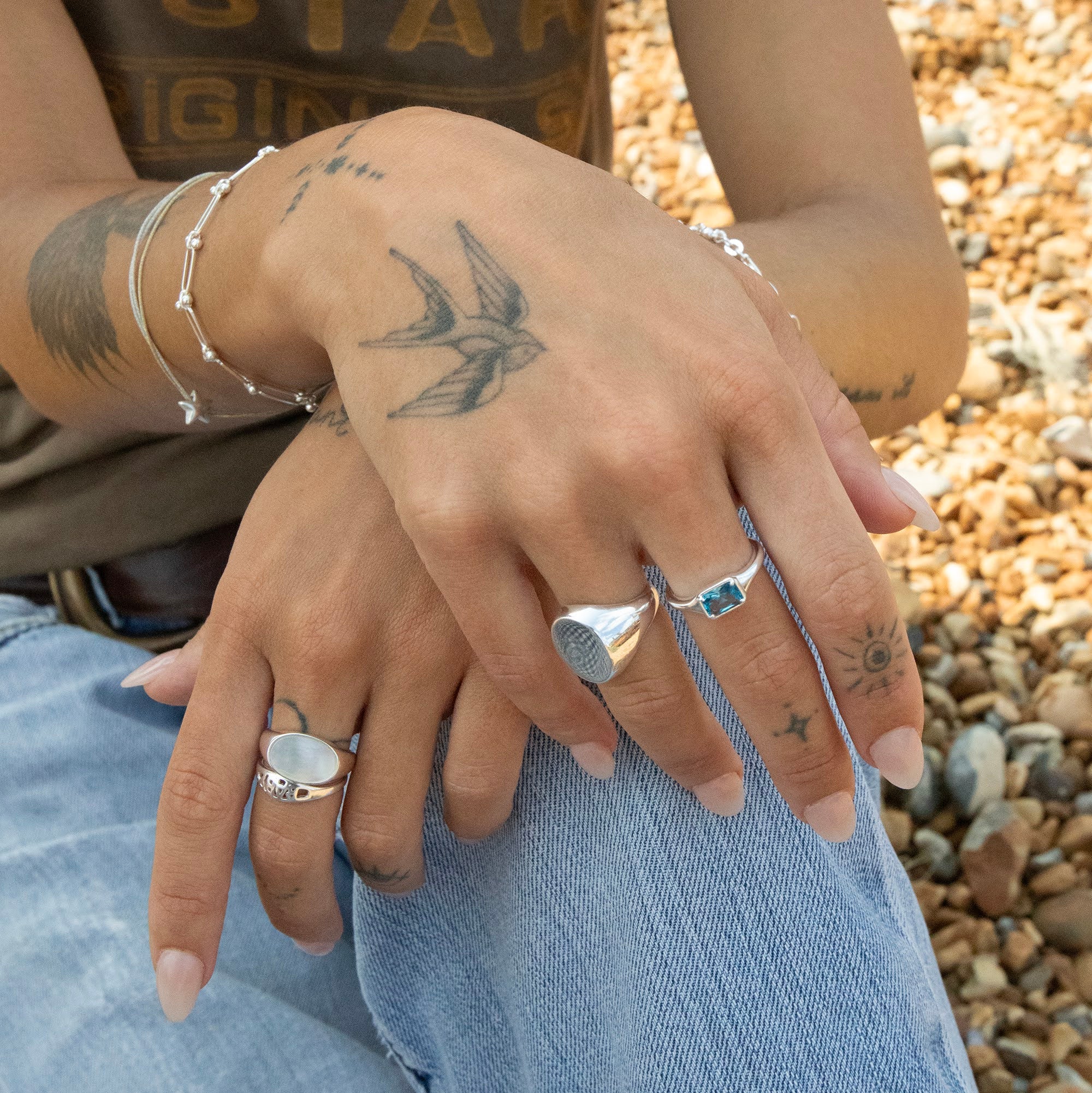 Close-up of hands with tattoos and rings on a pebbly surface - seolgold