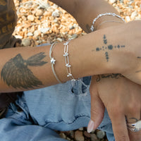 Close-up of a person's arm with silver bracelets and tattoos against a pebbly background - seolgold