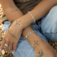 Close-up of a person's hands with tattoos and silver bracelets on a pebbly surface - seolgold