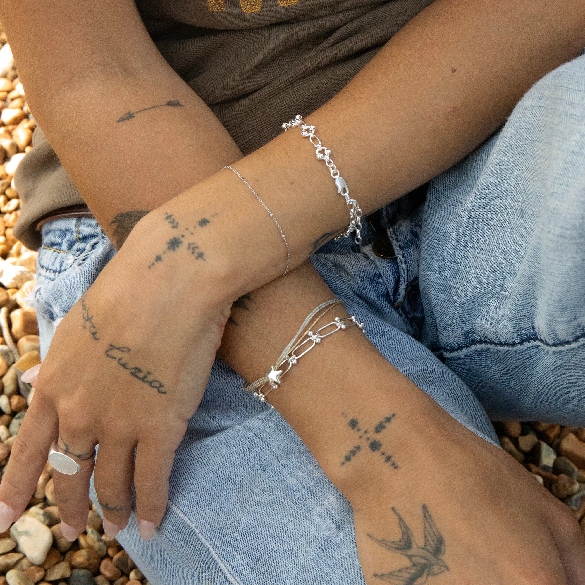 Close-up of a person's hands with tattoos and silver bracelets on a pebbly surface - seolgold