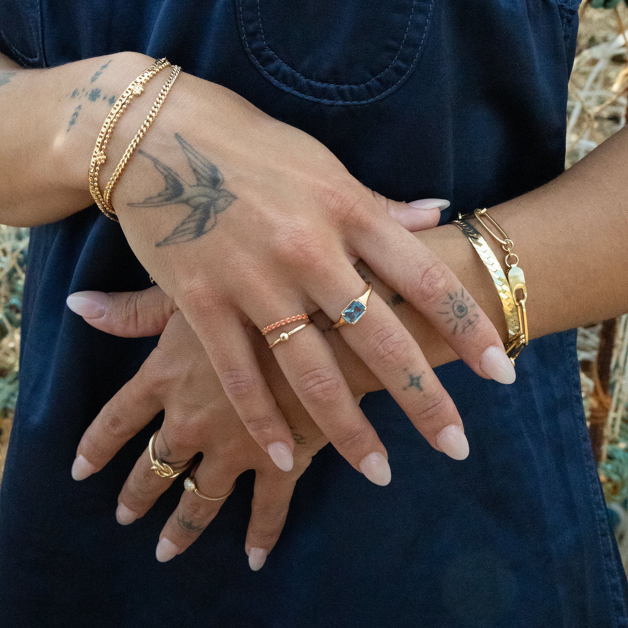 Close-up of hands with gold jewelry and tattoos against a dark blue fabric background - blue stone ring - seolgold