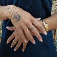 Close-up of hands with gold jewelry and tattoos against a dark blue fabric background - blue stone ring - seolgold