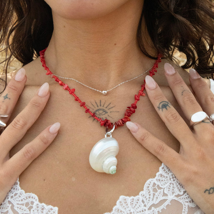 Person wearing a red beaded necklace with a shell pendant, surrounded by hands - seolgold