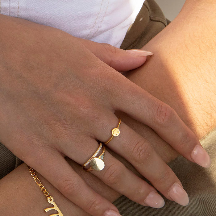 A close-up of a person's hands wearing gold rings, including a signet-style ring with a flat oval face and a thin ring with a small round charm, along with a gold chain bracelet - seolgold