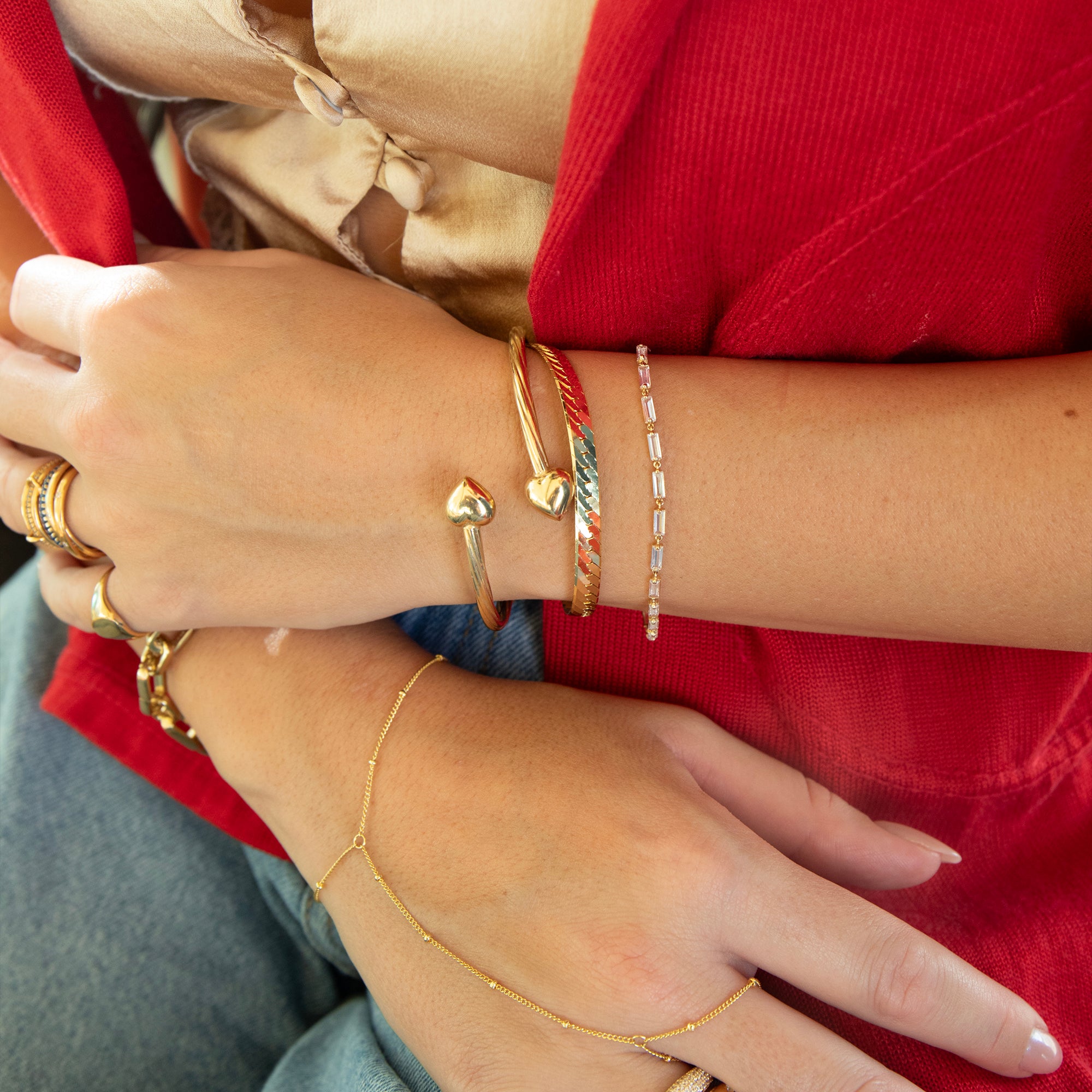 Close-up of hands wearing multiple gold bracelets and rings on a red background - seolgold