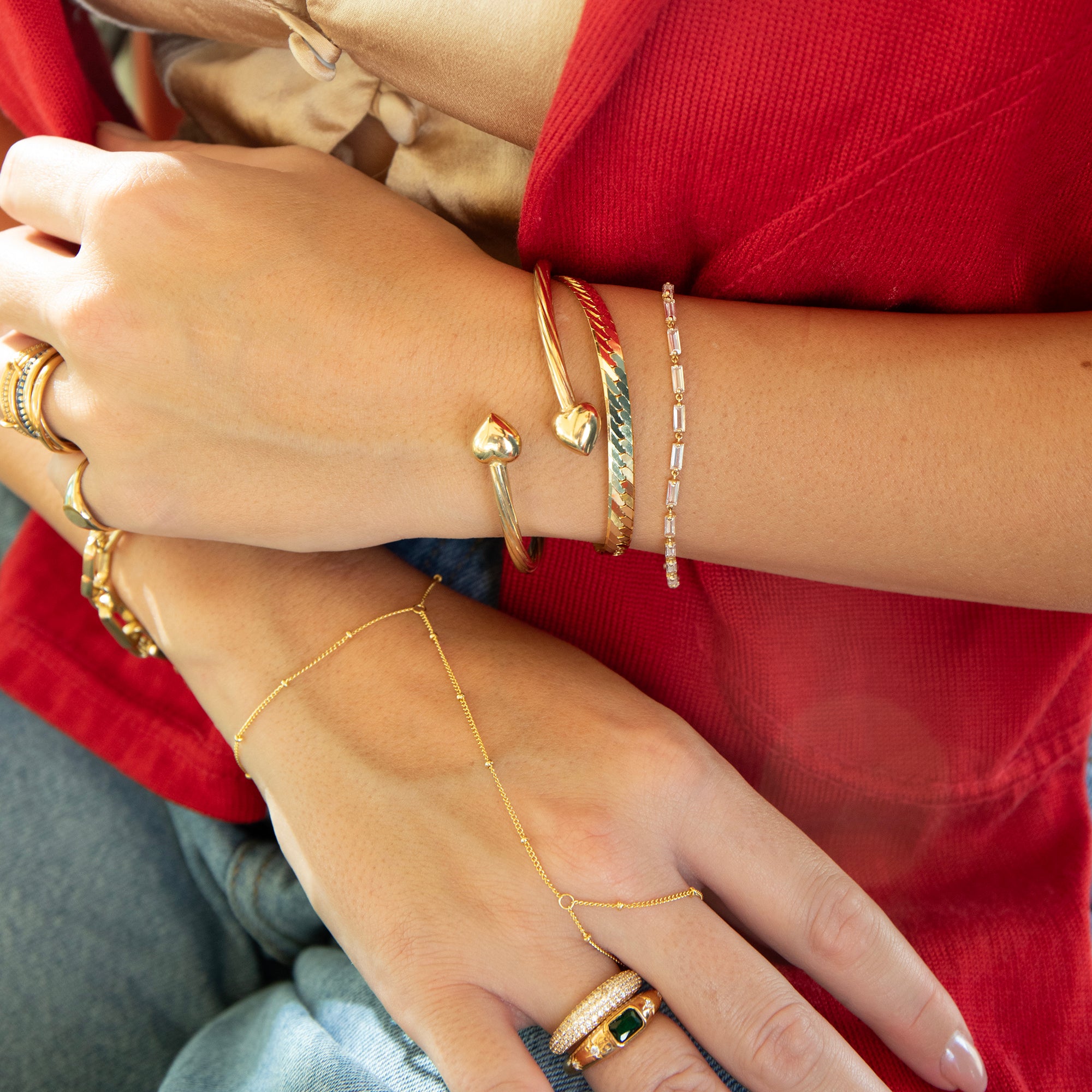 Close-up of hands wearing multiple gold bracelets and rings against a red background - seolgold