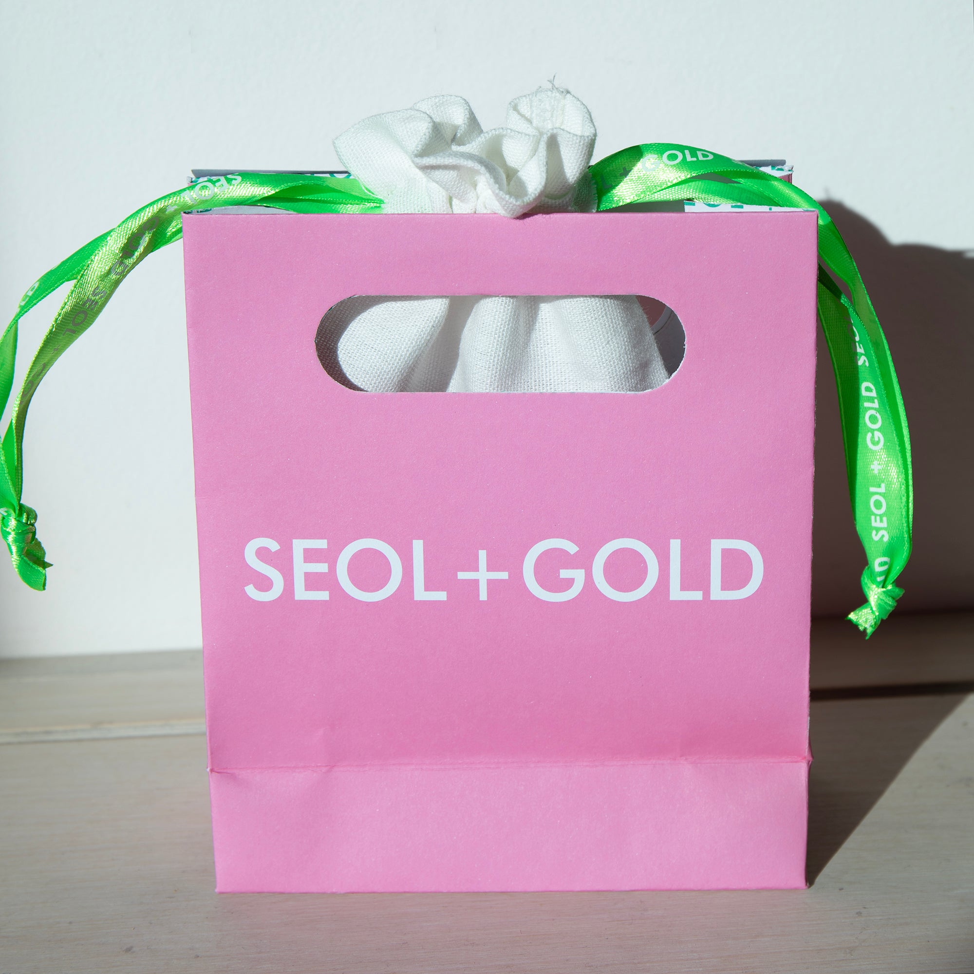 Pink gift bag with 'Seol+Gold' branding and a green ribbon on a light background - seolgold