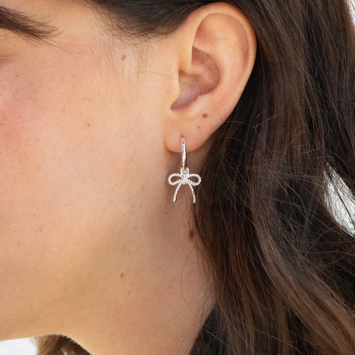 Close-up of an ear wearing a silver bow-shaped earring with dark hair and skin in the background - seolgold