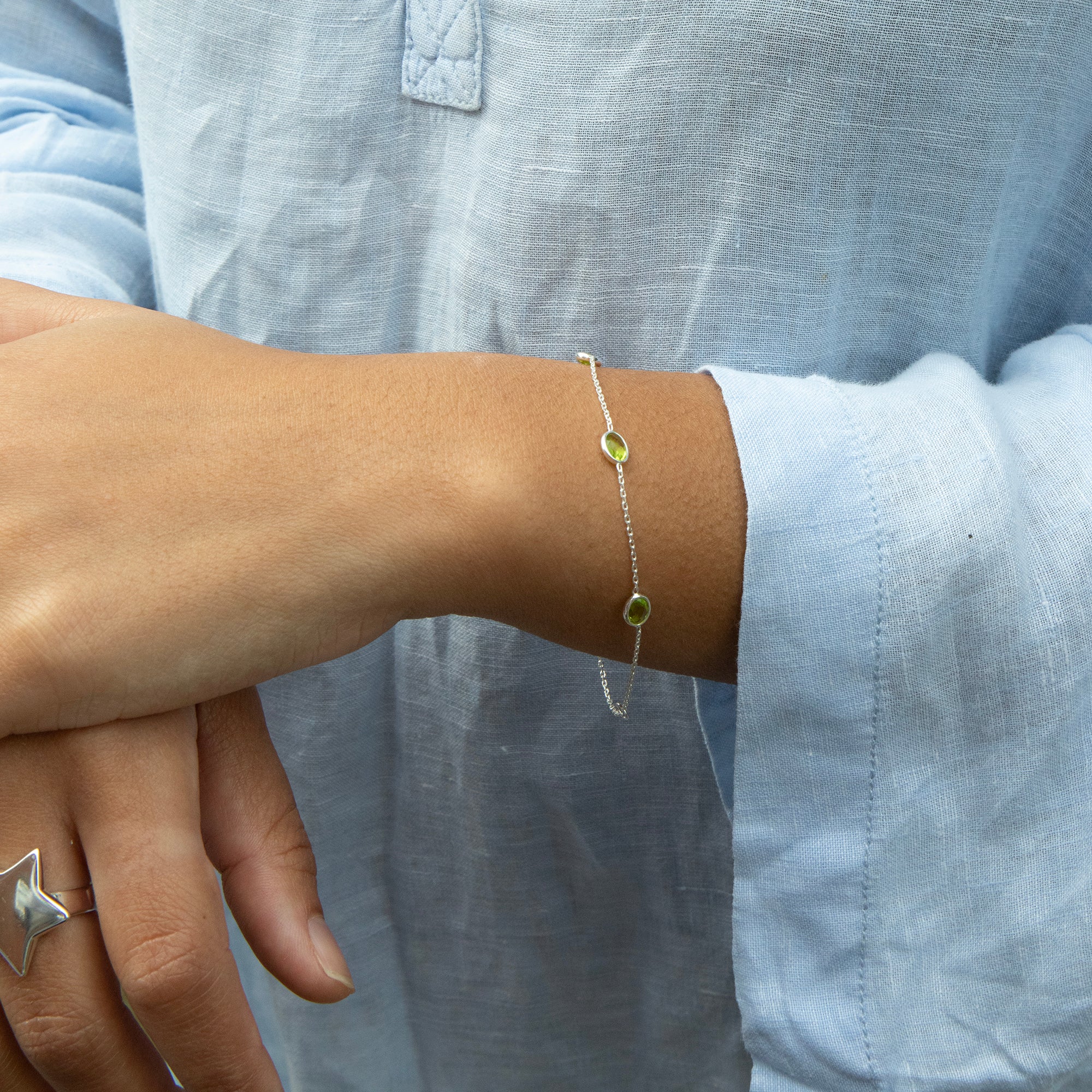 Close-up of a wrist wearing a bracelet with a green gemstone, against a light blue shirt background - seolgold