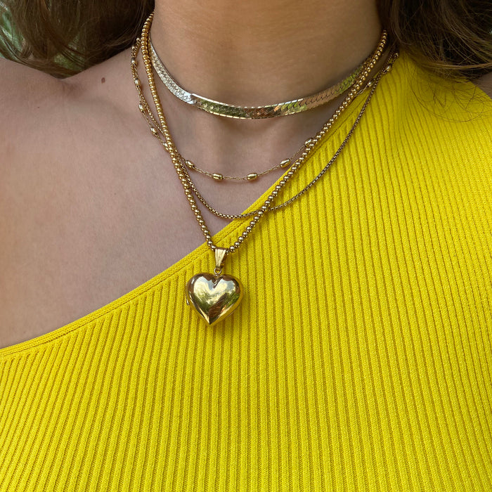 A close-up of a person wearing layered gold necklaces, including a heart-shaped pendant necklace, a beaded chain, and a textured choker, paired with a bright yellow ribbed top - seolgold