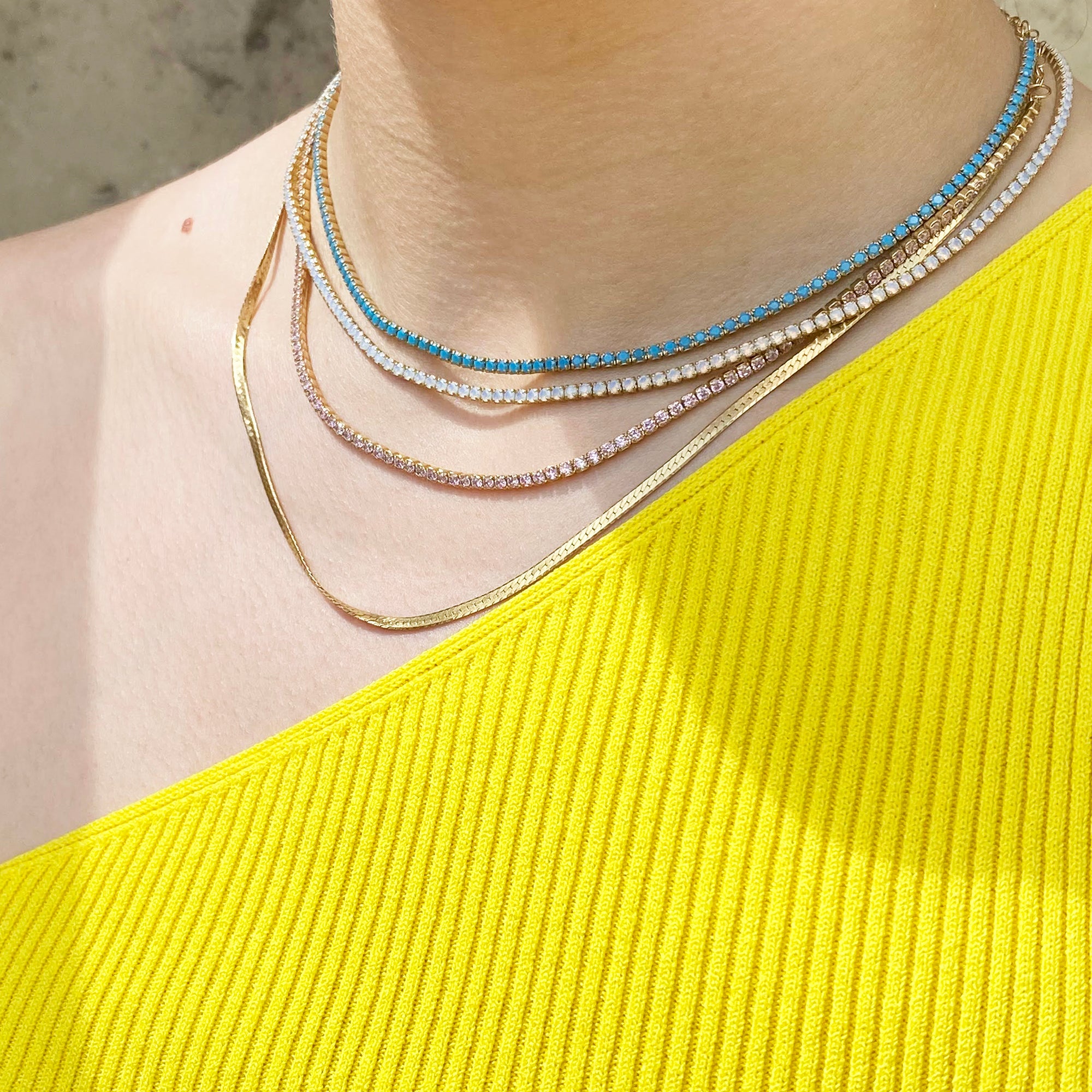 A close-up of a model's neck and upper chest wearing a layered necklace set. The set includes a gold snake chain, a gold chain with clear rhinestones, a gold chain with white rhinestones, and a delicate gold chain with light blue rhinestones - seolgold