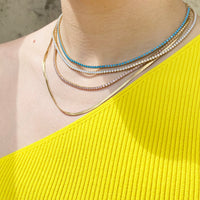 A close-up of a model's neck and upper chest wearing a layered necklace set. The set includes a gold snake chain, a gold chain with clear rhinestones, a gold chain with white rhinestones, and a delicate gold chain with light blue rhinestones - seolgold