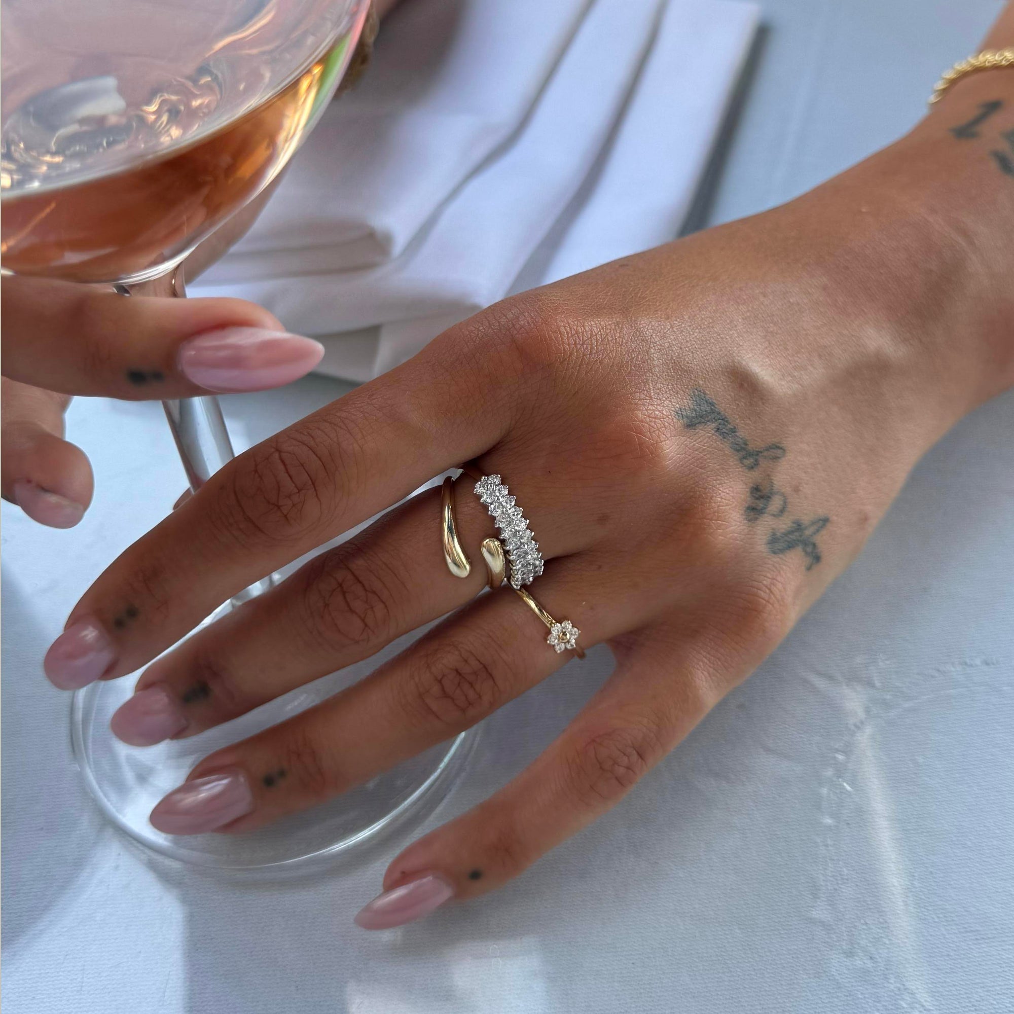 Hand with multiple rings holding a glass of rosé wine on a white surface - seolgold