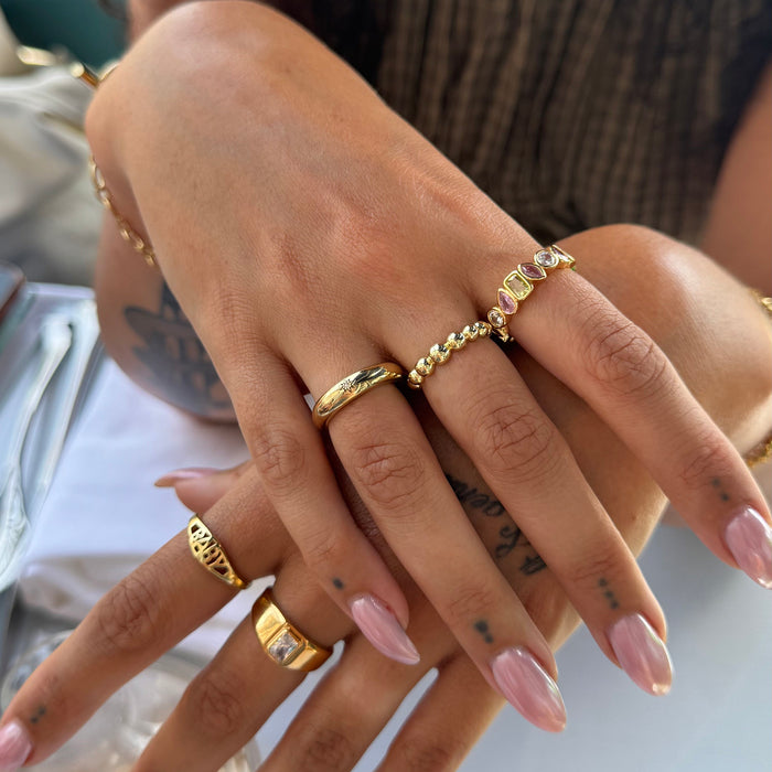 A close-up of a person's hands wearing multiple gold rings, including a beaded chain ring, a textured band ring, and a ring with a clear stone, against a softly blurred background - seolgold