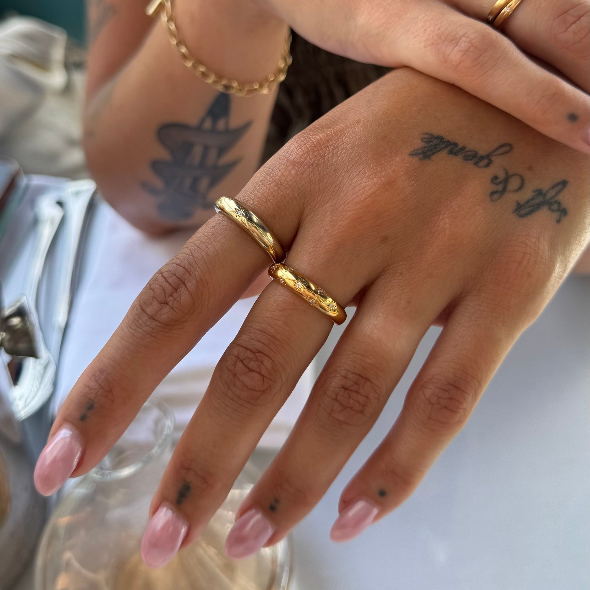 A close-up of a person's hand wearing two slim textured gold rings, with pink manicured nails and visible tattoos on the hand and forearm - seolgold