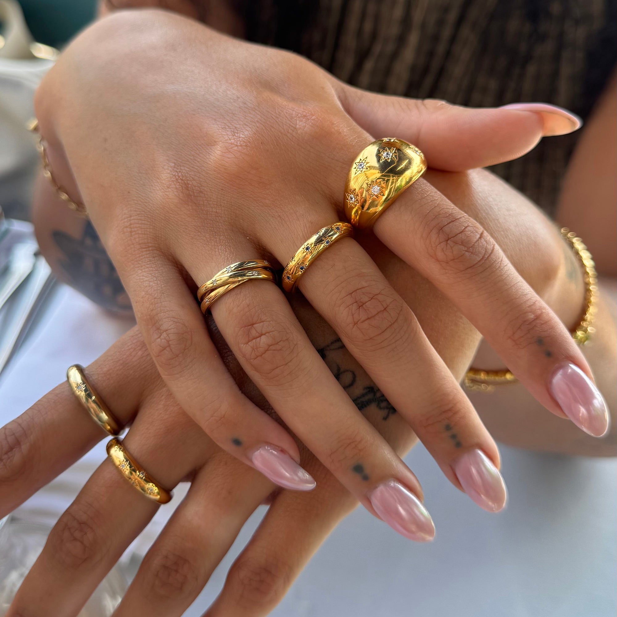 Close-up of a hand wearing multiple gold rings with pink nail polish on a blurred background - seolgold