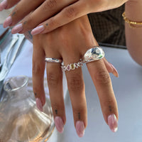 A close-up of a person's hand wearing a stack of silver rings, including a textured teardrop ring and a multi-colored stone band, with pale pink manicured nails - seolgold