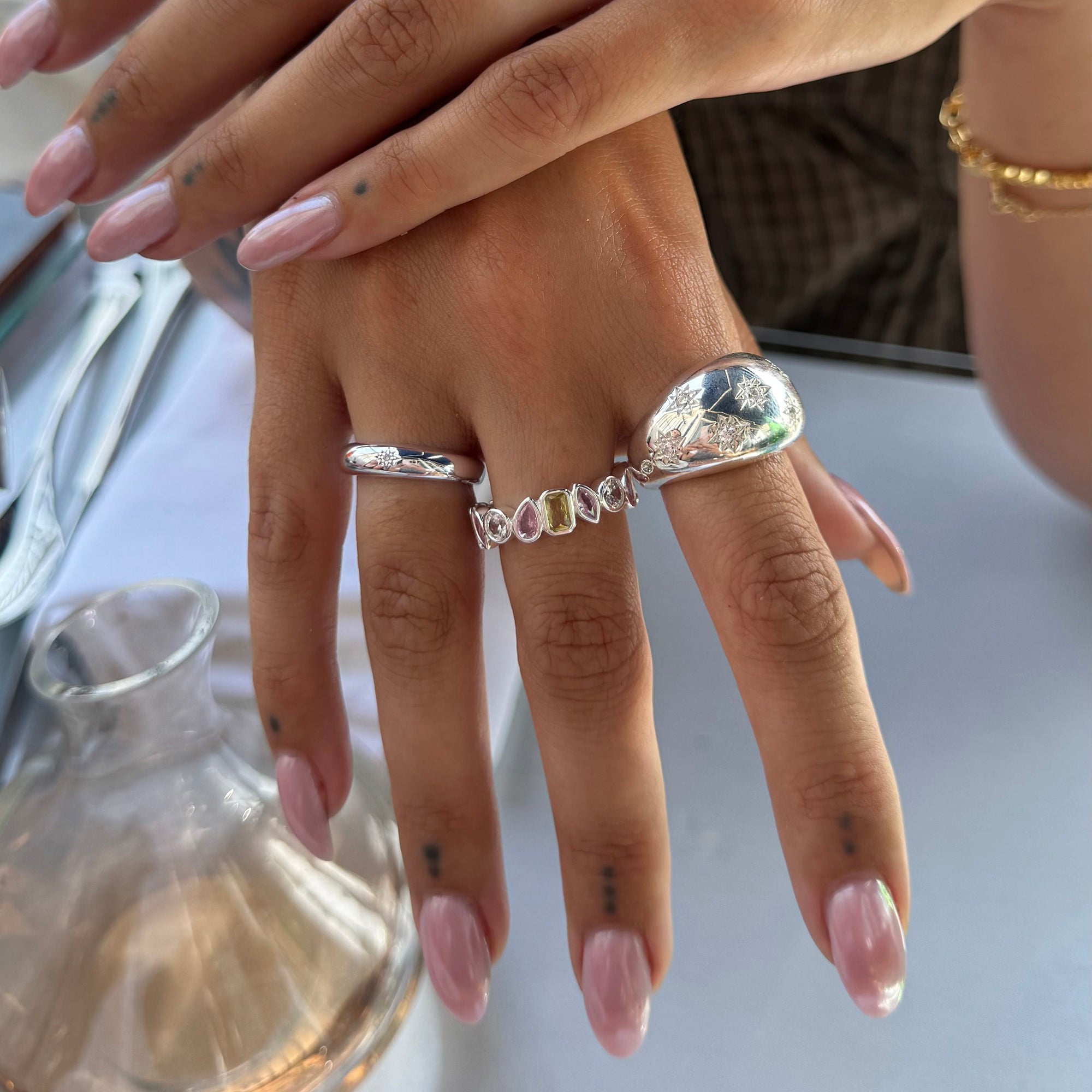 A close-up of a person's hand wearing a stack of silver rings, including a textured teardrop ring and a multi-colored stone band, with pale pink manicured nails - seolgold