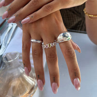 A close-up of a person's hand wearing a stack of silver rings, including a textured teardrop ring and a multi-colored stone band, with pale pink manicured nails - seolgold
