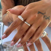 A close-up of a person's hand wearing multiple silver rings, including a textured band ring and a ring with a clear gemstone, against a softly blurred background - seolgold