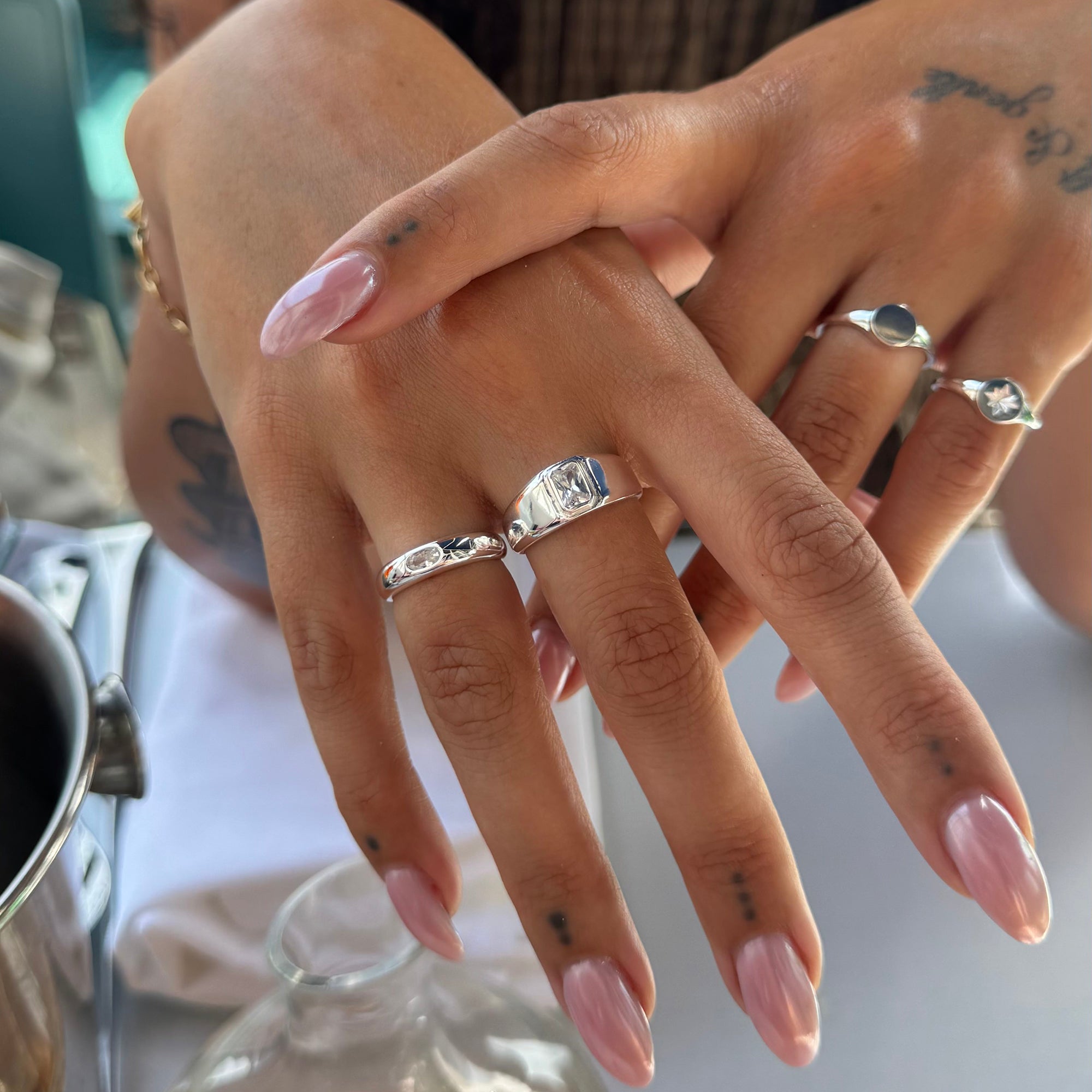 A close-up of a person's hands wearing multiple silver rings, including a wide band with a central rectangular stone and smaller rings with marquise-cut stones, against a softly blurred indoor background - seolgold