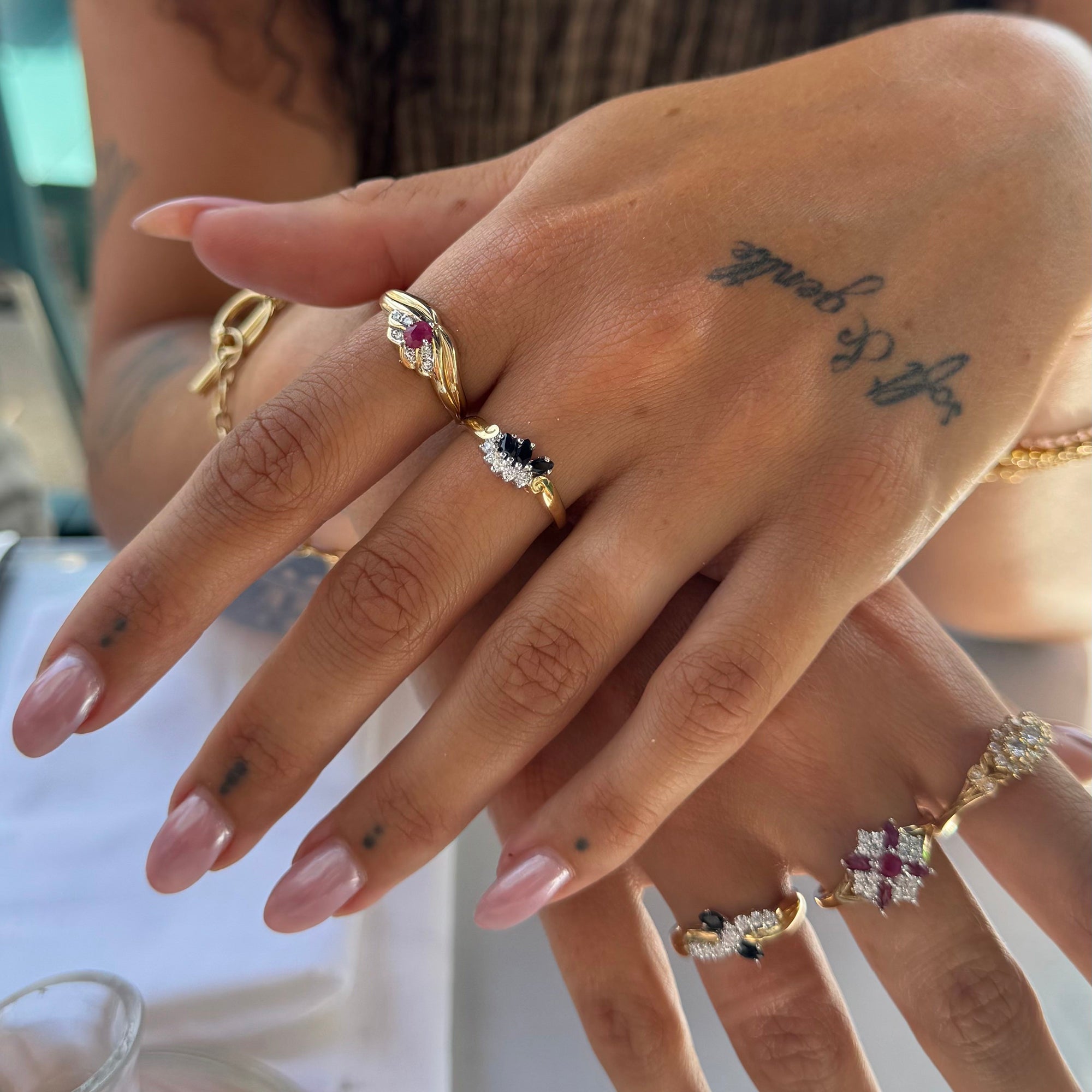 A close-up of a person's hands wearing multiple gold rings with colorful gemstones, including pink, black, and white stones - seolgold