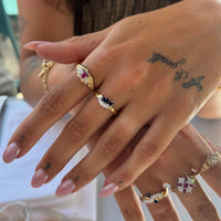 A close-up of a hand wearing multiple gold rings with colorful gemstones, including pink, blue, and clear stones, against a softly blurred background - seolgold