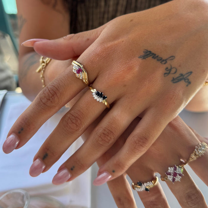 A close-up of a hand wearing multiple gold rings with colorful gemstones, including pink, blue, and clear stones, against a softly blurred background - seolgold