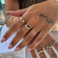 A close-up of a hand wearing multiple gold rings with colorful gemstones, including pink, blue, and clear stones, against a softly blurred background - seolgold