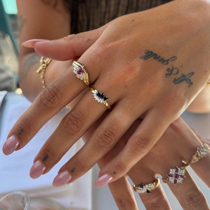 A close-up of a hand wearing multiple gold rings with colorful gemstones, including pink, blue, and clear stones, against a softly blurred background - seolgold