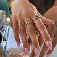 A close-up of a person's hands wearing multiple gold rings with blue and clear gemstones, set against a softly blurred cafe or indoor background - seolgold
